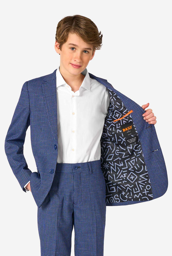 Een tienerjongen, gekleed in een marineblauw en blauw geruit pak met een wit overhemd. Het pak heeft een moderne pasvorm met twee knopen en klepzakken. Hij staat tegen een effen witte achtergrond. Houdt zijn jasje open