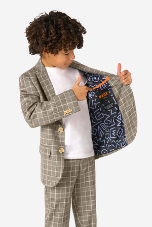 Een jongen, gekleed in een licht bruin geruit pak met een wit overhemd. Het pak heeft een moderne pasvorm met twee knopen en klepzakken. Hij staat tegen een effen witte achtergrond. Houdt een kant van het jasje open.