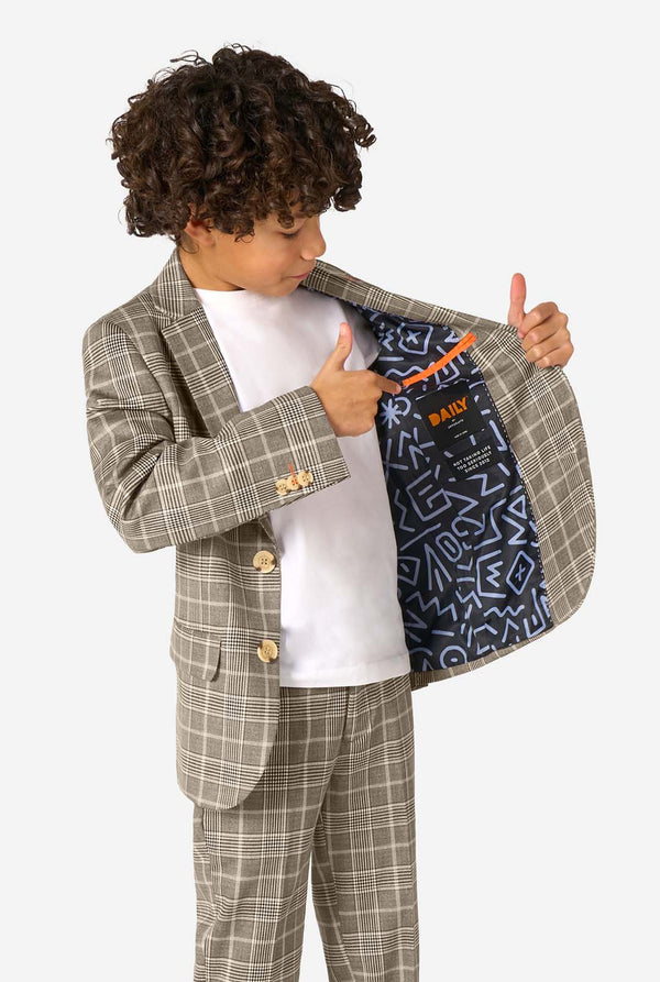 Een jongen, gekleed in een licht bruin geruit pak met een wit overhemd. Het pak heeft een moderne pasvorm met twee knopen en klepzakken. Hij staat tegen een effen witte achtergrond. Houdt een kant van het jasje open.
