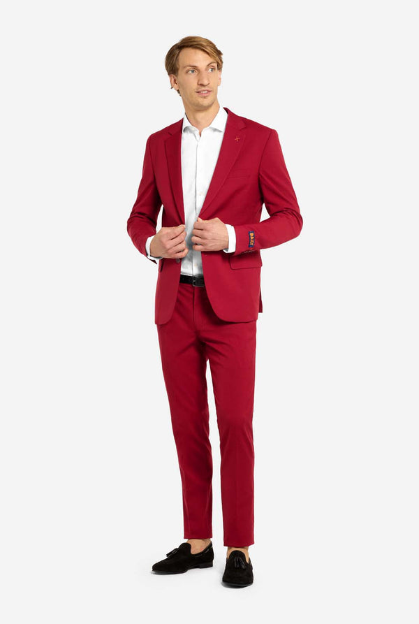 Afbeelding met lichtgrijze achtergrond en man die helder bordeauxrood mannenpak en wit overhemd draagt. Kostuum bestaat uit blazer en broek. Volledig lichaam.
