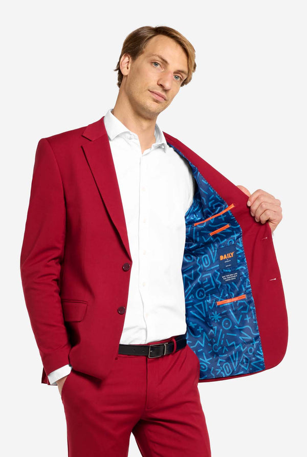 Afbeelding met lichtgrijze achtergrond en man die helder bordeauxrood mannenpak en wit overhemd draagt. Kostuum bestaat uit blazer en broek. Houd een kant van het jasje open.