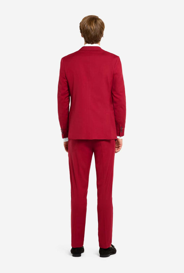 Afbeelding met lichtgrijze achtergrond en man die helder bordeauxrood mannenpak en wit overhemd draagt. Kostuum bestaat uit blazer en broek. Zicht vanaf de achterkant.