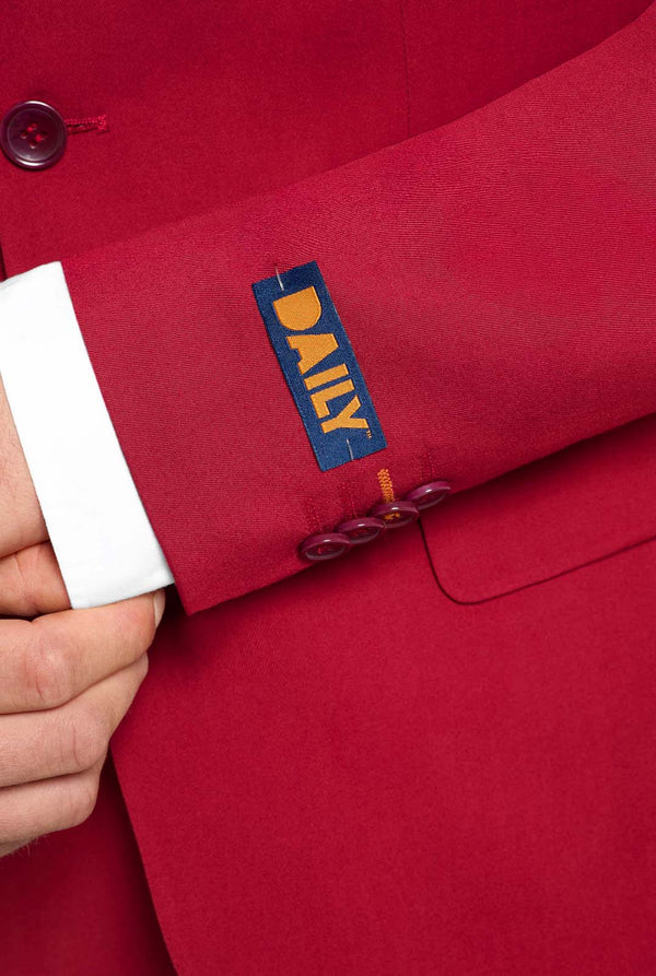 Afbeelding met lichtgrijze achtergrond en man die helder bordeauxrood mannenpak en wit overhemd draagt. Kostuum bestaat uit blazer en broek. Gericht op de mouwen.