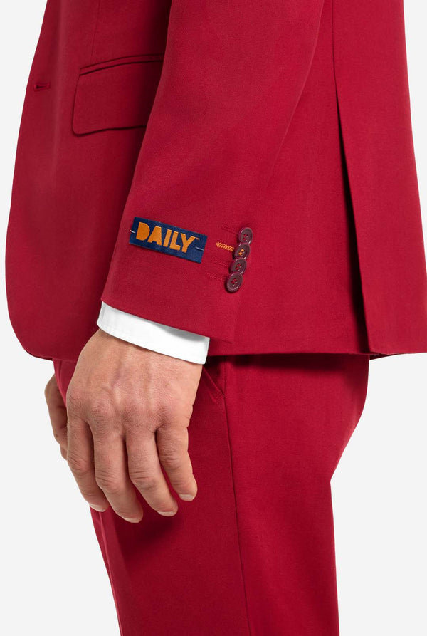 Afbeelding met lichtgrijze achtergrond en man die helder bordeauxrood mannenpak en wit overhemd draagt. Kostuum bestaat uit blazer en broek. Zicht vanaf de zijkant.