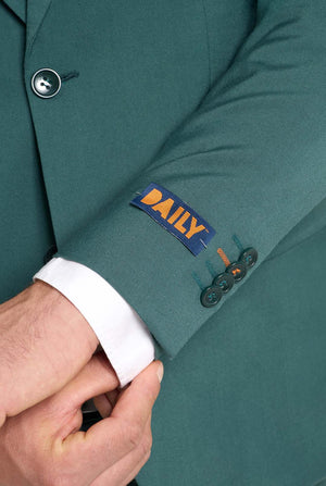 Afbeelding met lichtgrijze achtergrond en een man in een donkergroen herenpak en wit overhemd. Het pak bestaat uit een blazer en een pantalon. Close up van de mouwen