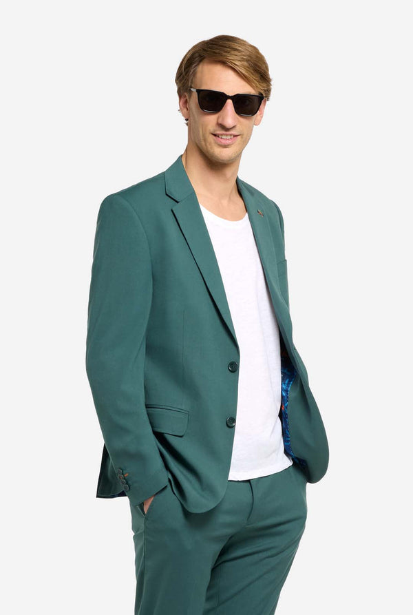 Afbeelding met lichtgrijze achtergrond en een man in een donkergroen herenpak en wit overhemd. Het pak bestaat uit een blazer en een pantalon. Kijkt naar de camera en draagt een zonnebril