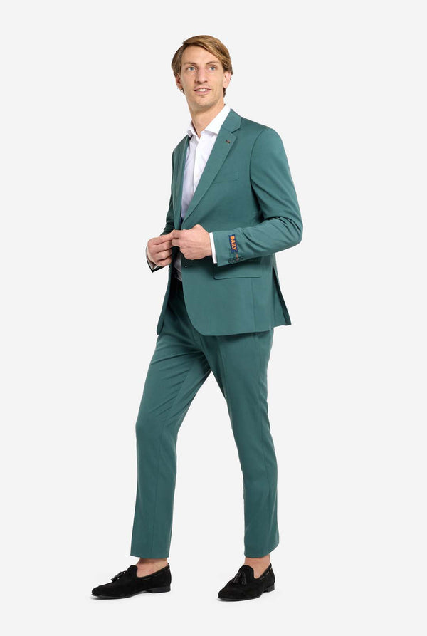 Afbeelding met lichtgrijze achtergrond en een man in een donkergroen herenpak en wit overhemd. Het pak bestaat uit een blazer en een pantalon. De man kijkt zijwaarts. Volledig lichaam foto