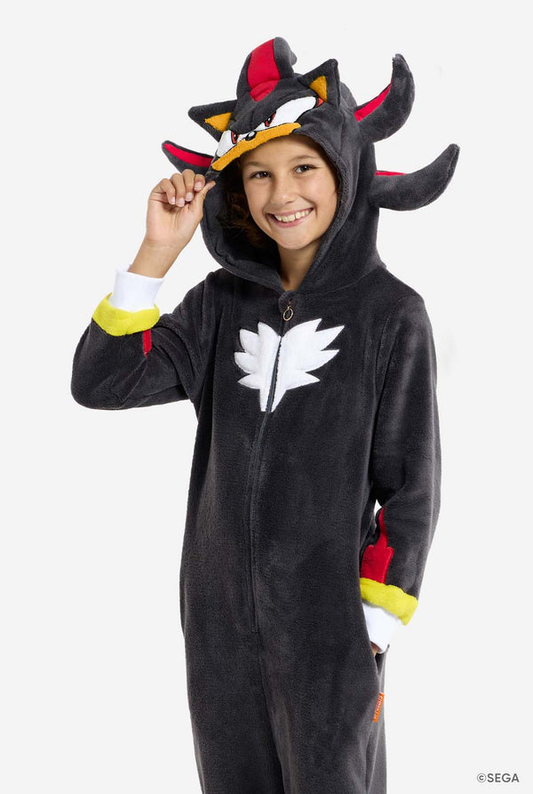 Een kind draagt ​​een pluchen, zwart kinderonesie, geïnspireerd op het populaire egelpersonage Shadow, met rode en zwarte accenten op de capuchon, een wit embleem op de borst en gele details op de polsen. De onesie is gemaakt van zacht fleecemateriaal en biedt comfort en een speels design voor cosplay, loungen of thema-evenementen. De persoon lacht met de capuchon op zijn hoofd. Perfect voor fans van games of animatiefiguren.