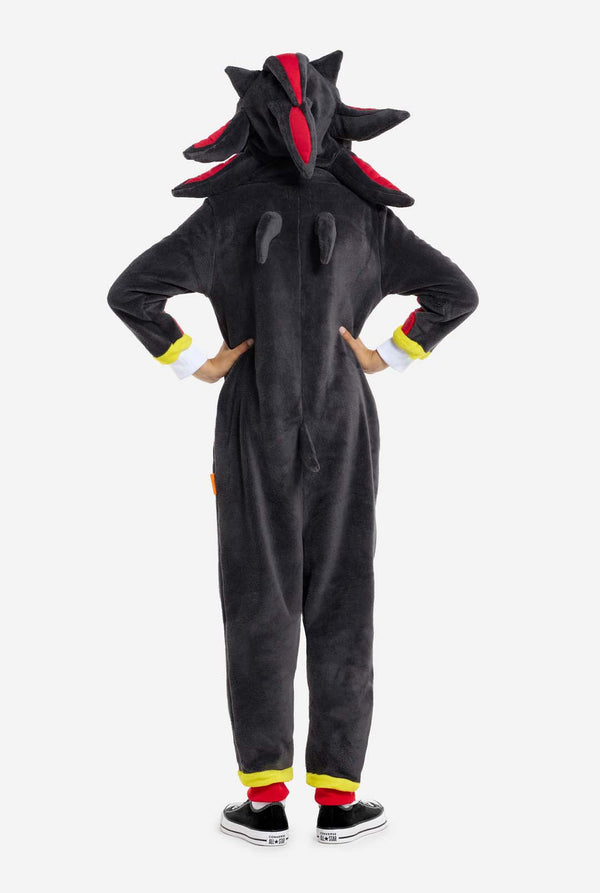Een kind dat een pluchen zwarte kinderonesie draagt, geïnspireerd op het populaire egelkarakter Shadow, met stekelige rode en zwarte accenten op de capuchon, een wit embleem op de borst en gele polsdetails. De onesie is gemaakt van zacht fleecemateriaal en biedt comfort en een speels ontwerp voor cosplay, loungen of thema-evenementen. Uitzicht vanaf de achterkant. Perfect voor fans van gaming of geanimeerde karakters.