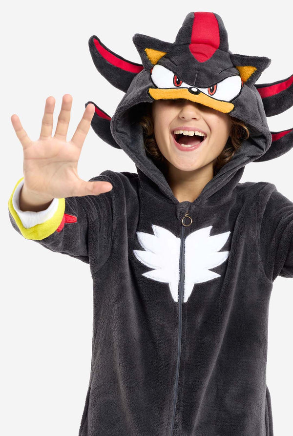 Een kind dat een pluchen zwarte kinderonesie draagt, geïnspireerd op het populaire egelkarakter Shadow, met stekelige rode en zwarte accenten op de capuchon, een wit embleem op de borst en gele polsdetails. De onesie is gemaakt van zacht fleecemateriaal en biedt comfort en een speels ontwerp voor cosplay, loungen of thema-evenementen. Close-up met de kap over de ogen. Perfect voor fans van gaming of geanimeerde karakters.