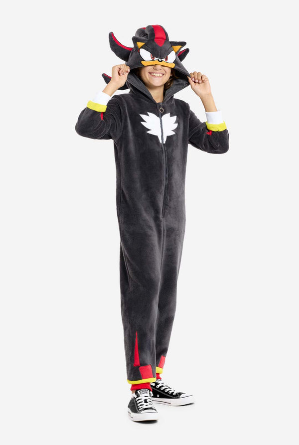 Een kind dat een pluchen zwarte kinderonesie draagt, geïnspireerd op het populaire egelkarakter Shadow, met stekelige rode en zwarte accenten op de capuchon, een wit embleem op de borst en gele polsdetails. De onesie is gemaakt van zacht fleecemateriaal en biedt comfort en een speels ontwerp voor cosplay, loungen of thema-evenementen. De persoon glimlacht terwijl hij de capuchon aanpast, wat het herkenbare gezicht en het opvallende ontwerp van het personage laat zien.