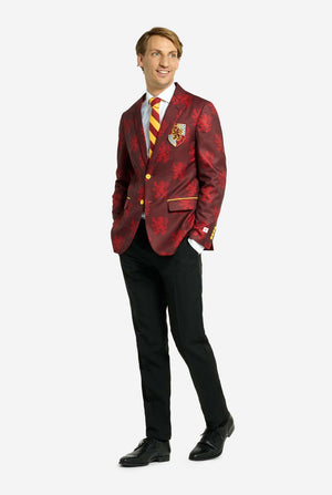 Afbeelding met lichtgrijze achtergrond en een man in een Harry Potter-pak in Griffoendor-stijl. Het pak bestaat uit een rode blazer met Griffoendor-logo en lichtrode leeuwenprint, een zwarte broek en een rood-gele diagonaal gestreepte stropdas. De man houdt zijn handen in zijn zakken en kijkt zijwaarts.