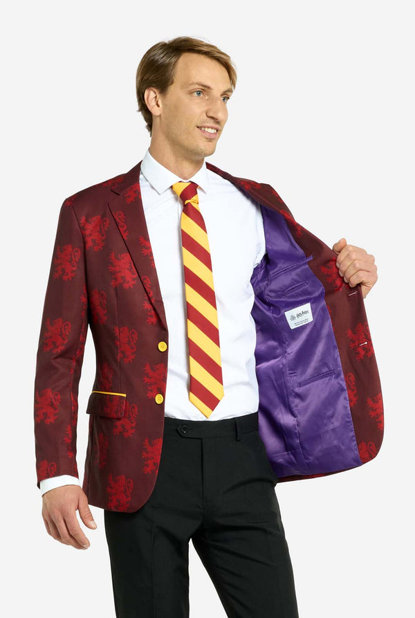 Afbeelding met lichtgrijze achtergrond en een man in een Harry Potter-pak in Griffoendor-stijl. Het pak bestaat uit een rode blazer met Griffoendor-logo en lichtrode leeuwenprint, een zwarte broek en een rood-gele diagonaal gestreepte stropdas. De man houdt één hand van de blazer open, waardoor de paarse binnenkant zichtbaar is.