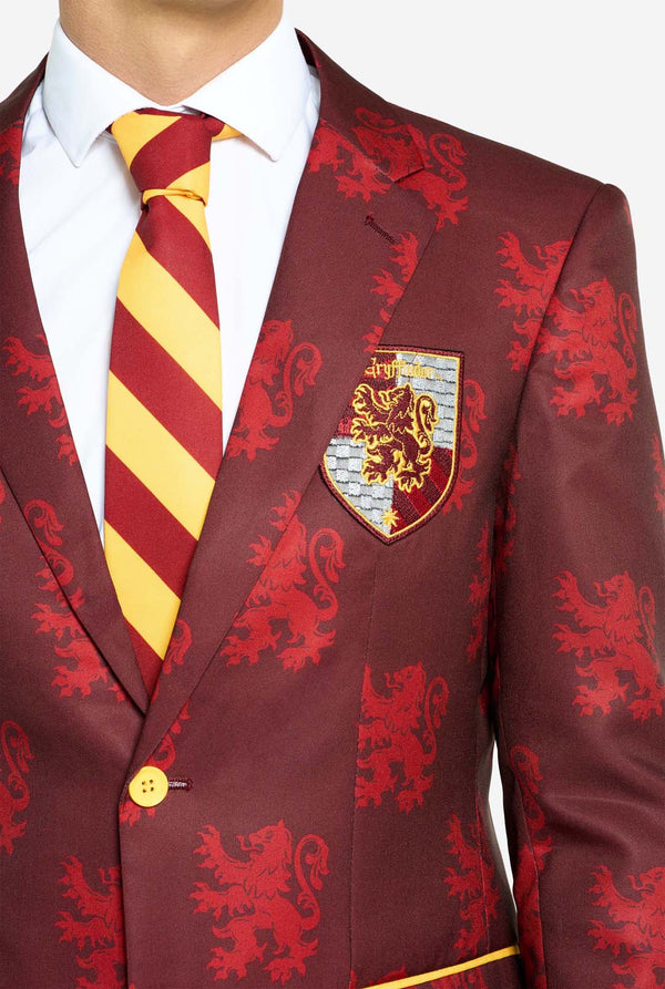 Afbeelding met lichtgrijze achtergrond en een man in een Harry Potter-pak in Griffoendor-stijl. Het pak bestaat uit een rode blazer met Griffoendor-logo en lichtrode leeuwenprint, een zwarte broek en een rood-gele diagonaal gestreepte stropdas. Close-up van de borst.