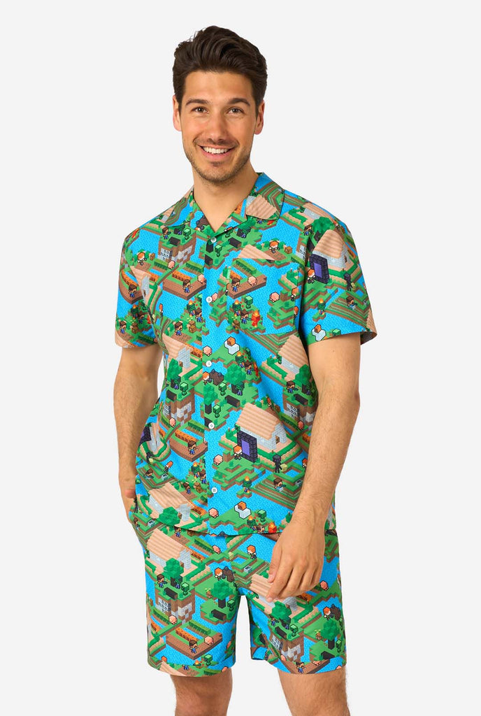 Een man draagt een kleurrijke zomeroutfit met een Minecraft Overworld print, met pixelachtige landschappen, personages en elementen uit het iconische spel. Het shirt met korte mouwen en de bijpassende shorts benadrukken een levendig ontwerp geïnspireerd door het Minecraft-universum.