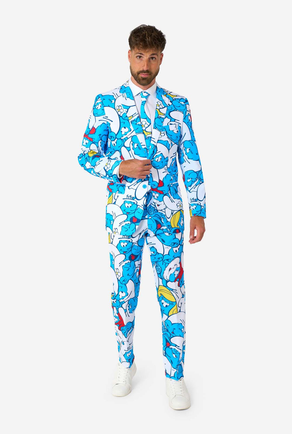 Lachende man gekleed in een kleurrijk herenpak van de Smurfen met een all-over cartoonprint in de kleuren blauw, wit, rood en geel, inclusief bijpassende stropdas en wit overhemd, staande tegen een effen achtergrond.