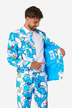 Lachende man gekleed in een kleurrijk herenpak van de Smurfen met een all-over cartoonprint in de kleuren blauw, wit, rood en geel, inclusief bijpassende stropdas en wit overhemd, staande tegen een effen achtergrond. Houd een kant van het jasje open.