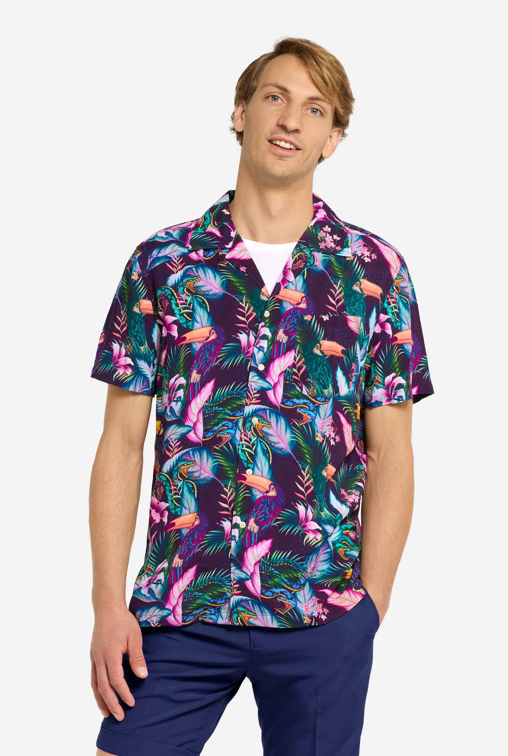 Een man draagt ​​een paars Hawaiiaans shirt met een tropische print, die doet denken aan het nachtleven in de jungle. De print bestaat uit papegaaien, slangen, bladeren en bloemen. Hij draagt ​​ook een paarse korte broek. Hij lacht in de camera en houdt één hand in zijn zak.