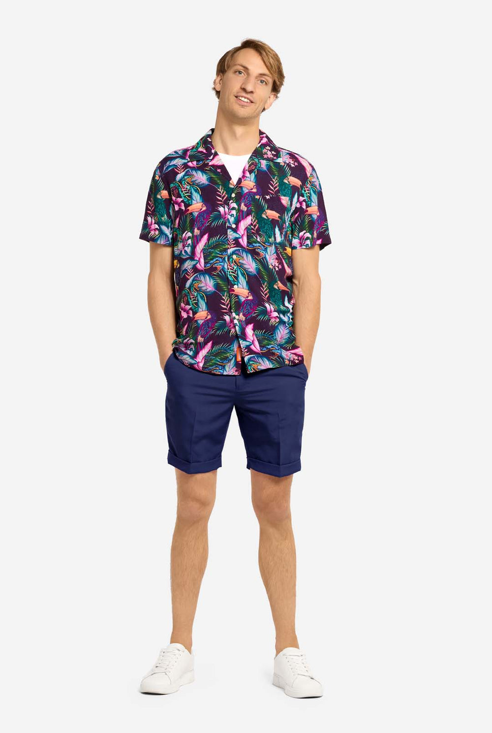 Een man draagt ​​een paars Hawaiiaans shirt met een tropische print, die doet denken aan het nachtleven in de jungle. De print bestaat uit papegaaien, slangen, bladeren en bloemen. Hij draagt ​​ook een paarse korte broek. Hij lacht in de camera en houdt één hand in zijn zak.