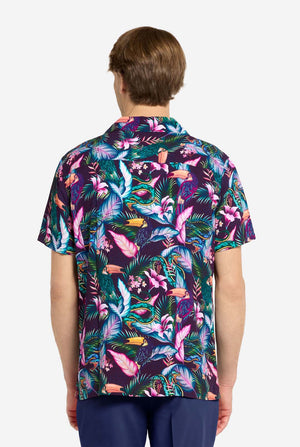 Een man draagt ​​een paars Hawaiiaans shirt met een tropische print, die doet denken aan het nachtleven in de jungle. De print bestaat uit papegaaien, slangen, bladeren en bloemen. Hij draagt ​​ook een paarse korte broek. Hij lacht in de camera en houdt één hand in zijn zak. Foto vanaf de achterkant.