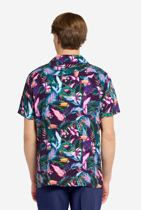 Een man draagt ​​een paars Hawaiiaans shirt met een tropische print, die doet denken aan het nachtleven in de jungle. De print bestaat uit papegaaien, slangen, bladeren en bloemen. Hij draagt ​​ook een paarse korte broek. Hij lacht in de camera en houdt één hand in zijn zak. Foto vanaf de achterkant.