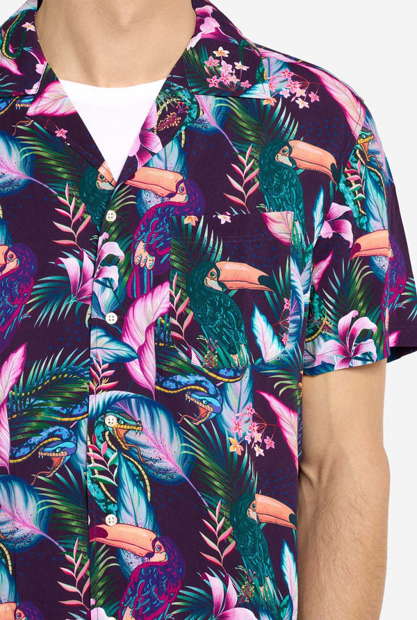 Een man draagt ​​een paars Hawaiishirt met een tropische print, dat doet denken aan het jungle-nachtleven. Print bestaat uit papegaaien, slangen, bladeren en bloemen. Draag ook een roze korte broek. Hij lacht in de camera en houdt één hand in de zak. Close-up.