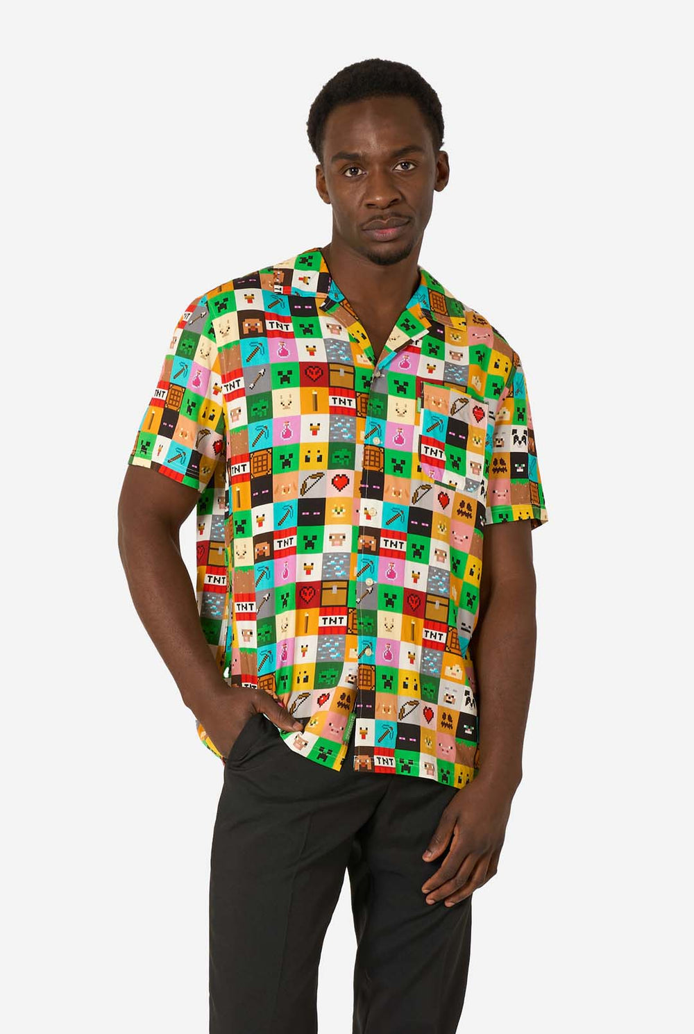 Man met een kleurrijk Minecraft Hawaiiaans shirt met gepixelde blokken, personages en items uit de game, gecombineerd met een zwarte broek, staande tegen een effen witte achtergrond.
