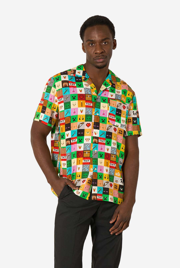 Man met een kleurrijk Minecraft Hawaiiaans shirt met gepixelde blokken, personages en items uit de game, gecombineerd met een zwarte broek, staande tegen een effen witte achtergrond.