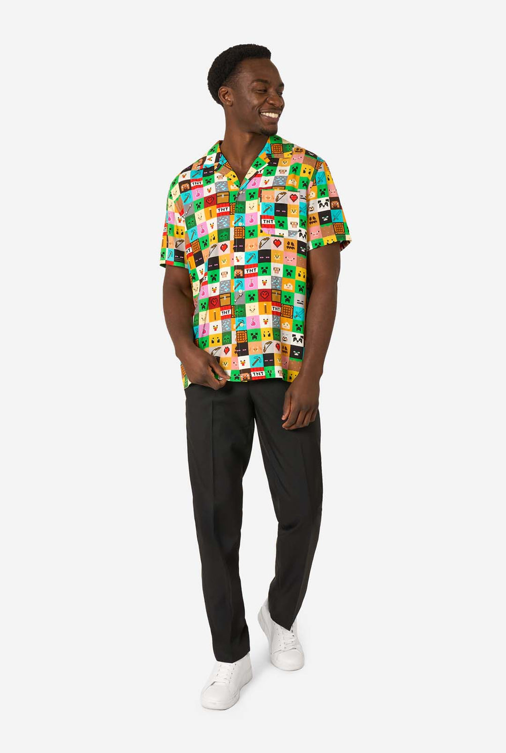 Man met een kleurrijk Minecraft Hawaiiaans shirt met gepixelde blokken, personages en items uit de game, gecombineerd met een zwarte broek, staande tegen een effen witte achtergrond.