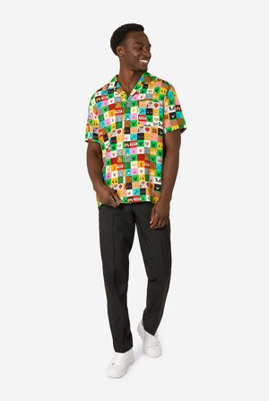 Man met een kleurrijk Minecraft Hawaiiaans shirt met gepixelde blokken, personages en items uit de game, gecombineerd met een zwarte broek, staande tegen een effen witte achtergrond. Volledig lichaam