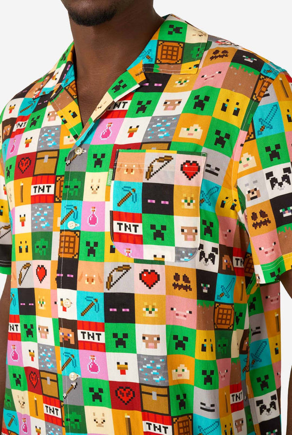 Man met een kleurrijk Minecraft Hawaiiaans shirt met gepixelde blokken, personages en items uit de game, gecombineerd met een zwarte broek, staande tegen een effen witte achtergrond. Close up van de borst