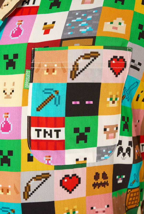 Man met een kleurrijk Minecraft Hawaiiaans shirt met gepixelde blokken, personages en items uit de game, gecombineerd met een zwarte broek, staande tegen een effen witte achtergrond. Close up van het borstzakje