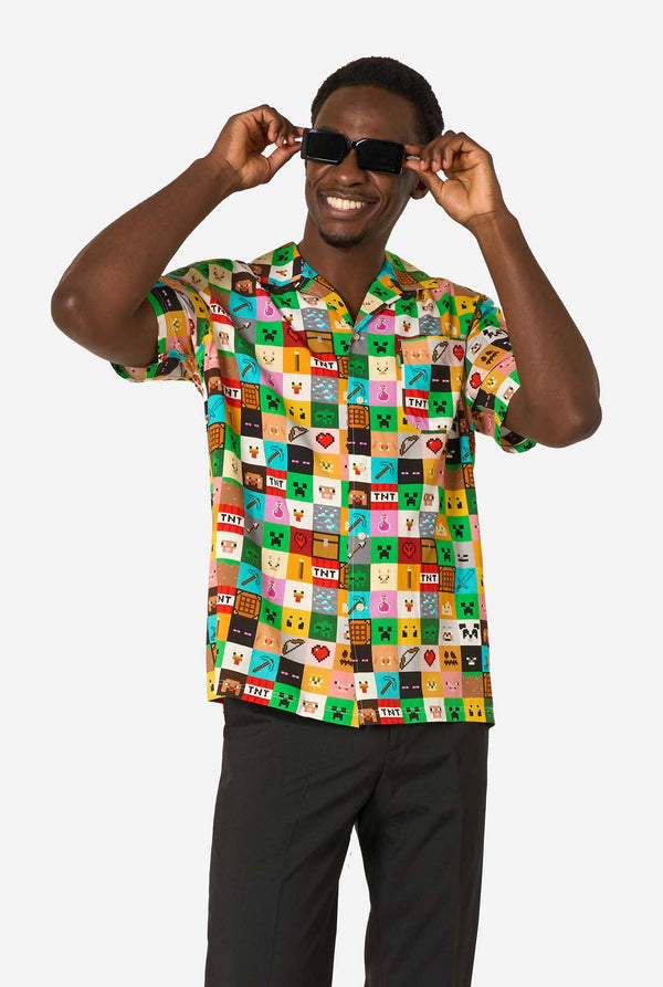 Man met een kleurrijk Minecraft Hawaiiaans shirt met gepixelde blokken, personages en items uit de game, gecombineerd met een zwarte broek, staande tegen een effen witte achtergrond. 