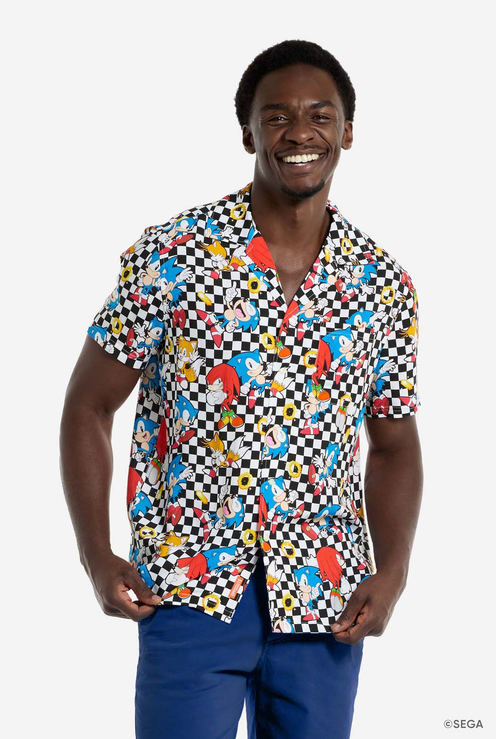 Een man draagt een zwart-wit geruit Hawaï-shirt met personages uit de Sonic the Hedgehog-franchise en gouden ringen erop. Hij lacht in de camera.