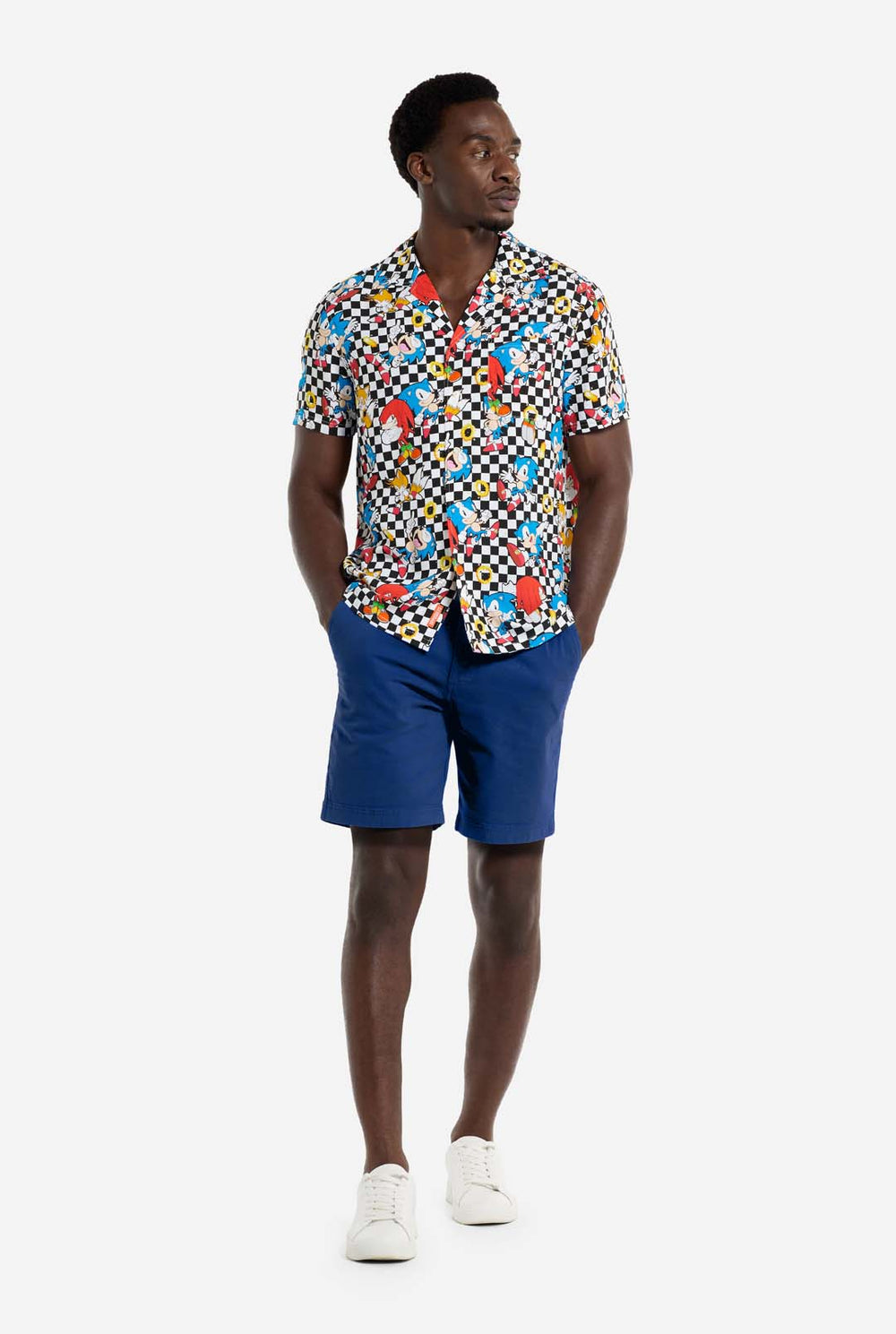 Een man draagt een zwart-wit geruit Hawaï-shirt met personages uit de Sonic the Hedgehog-franchise en gouden ringen erop. Hij lacht in de camera.