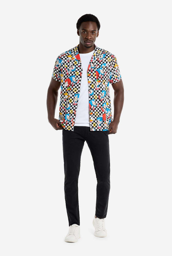 Een man draagt ​​een zwart-wit geruit Hawaï-shirt met personages uit de Sonic the Hedgehog-franchise en gouden ringen erop. Draag ook een zwarte broek. Hij kijkt in de camera en houdt de onderkant van het shirt vast.