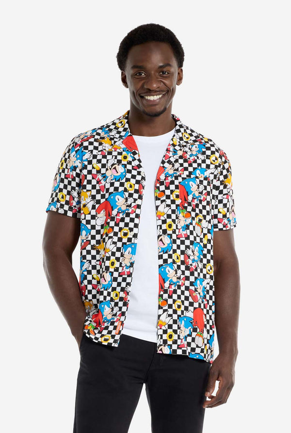 Een man draagt ​​een zwart-wit geruit Hawaï-shirt met personages uit de Sonic the Hedgehog-franchise en gouden ringen erop. Draag ook een zwarte broek. Hij lacht in de camera.