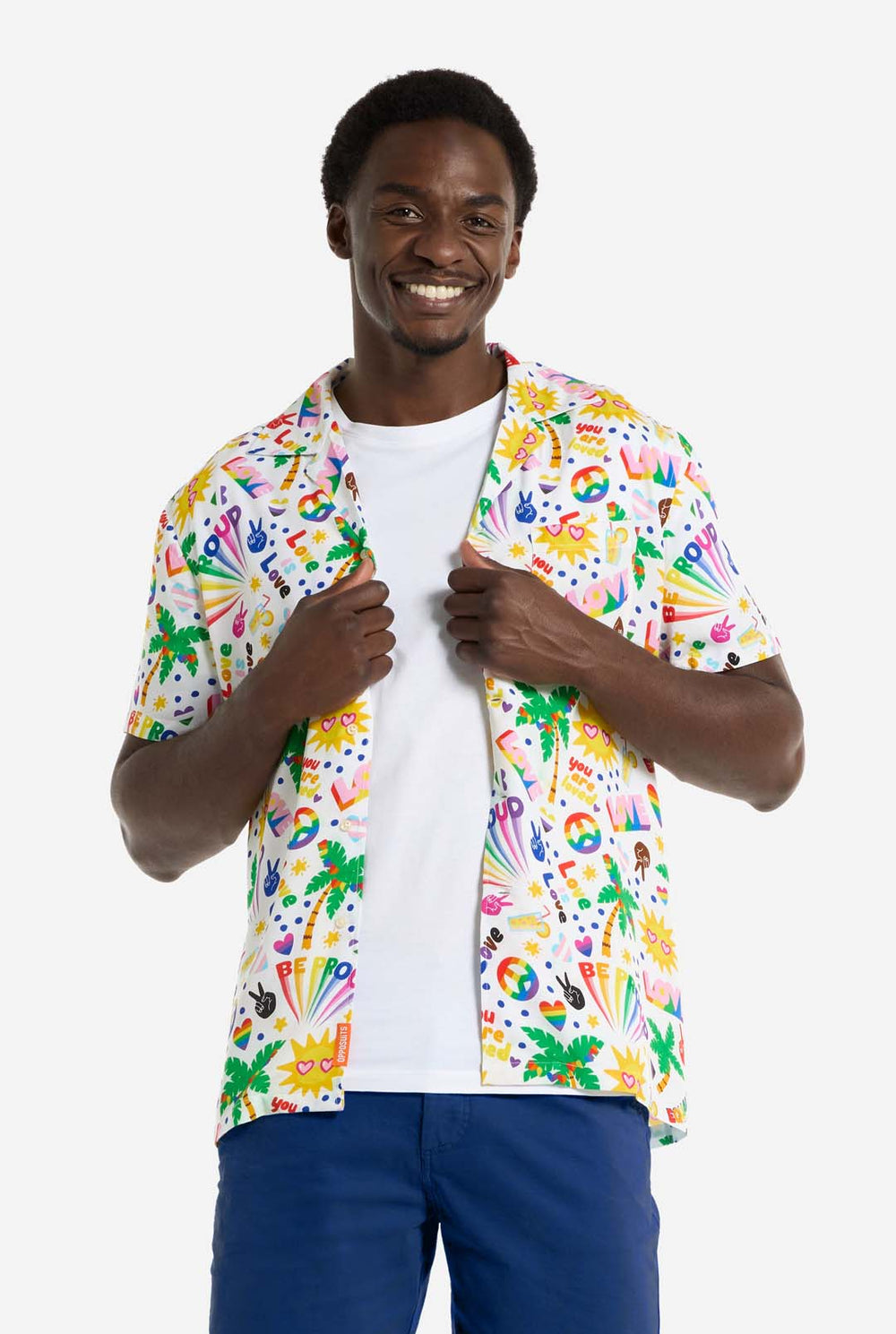 Een man draagt ​​een wit Hawaiiaans shirt met een regenboogprint. De print bestaat uit vredessymbolen, hartjes en andere iconen. Hij draagt ​​ook een blauwe broek. Hij lacht naar de camera en houdt de zijkanten van het shirt vast.