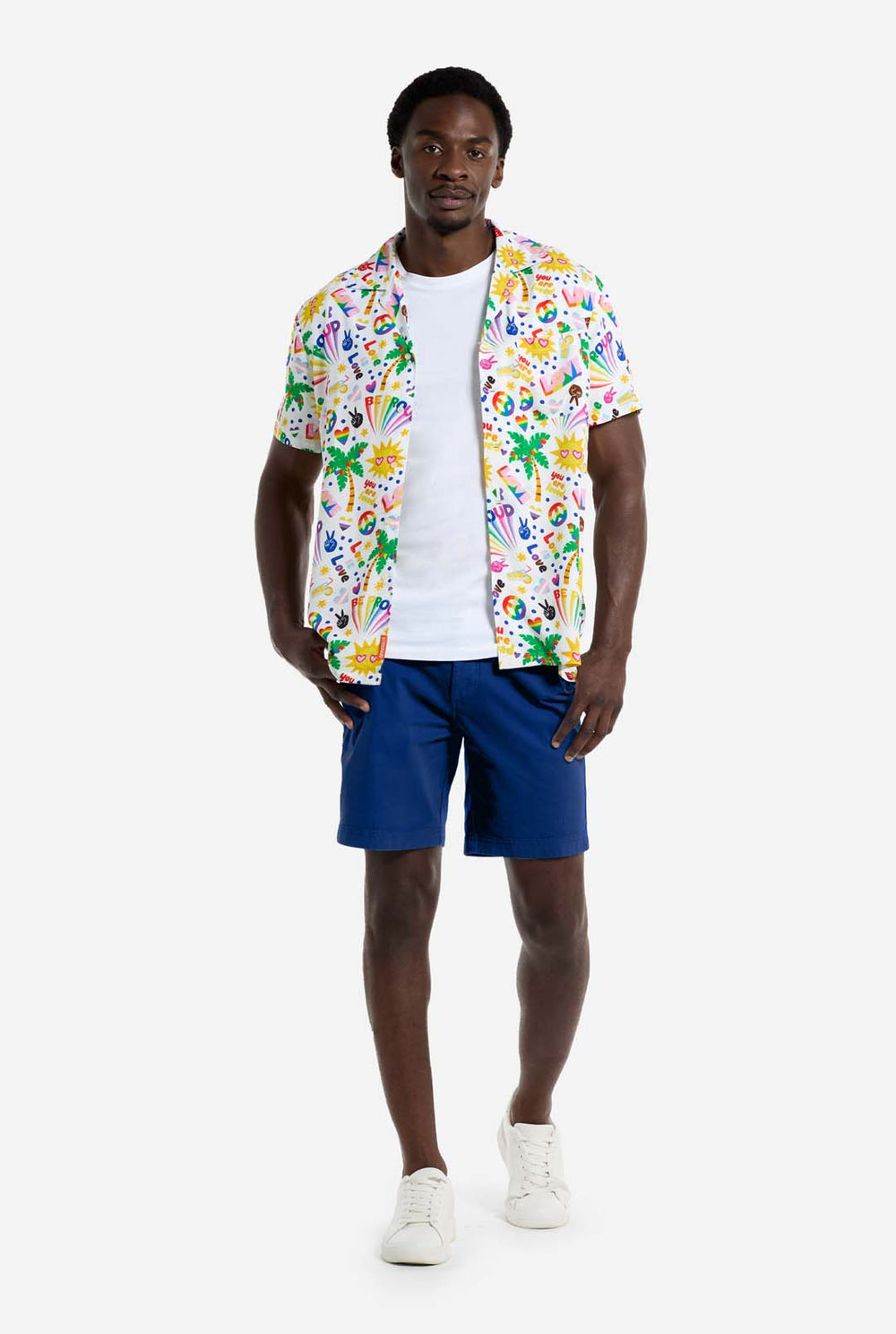Een man draagt ​​een wit Hawaiiaans shirt met een regenboogprint. De print bestaat uit vredessymbolen, hartjes en andere iconen. Hij draagt ​​ook een blauwe broek. Hij lacht naar de camera en houdt de zijkanten van het shirt vast.