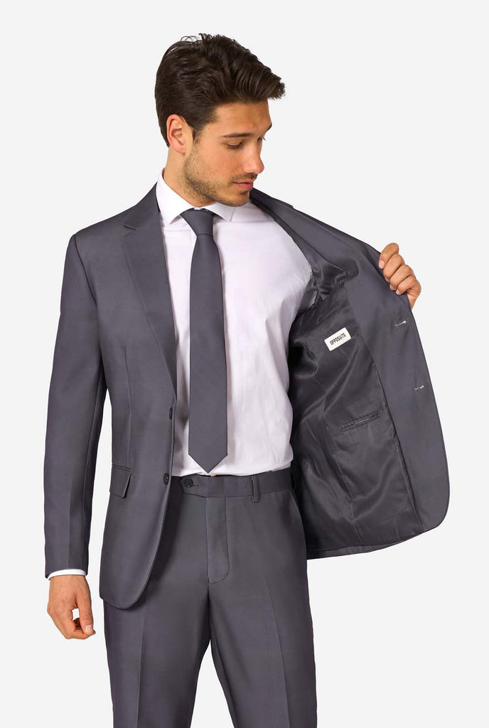 Een man draagt ​​een stijlvol antracietgrijs pak met een wit overhemd en ziet er zelfverzekerd uit. Perfecte formele outfit voor speciale gelegenheden, bruiloften en feesten. 1 kant van jasje open.