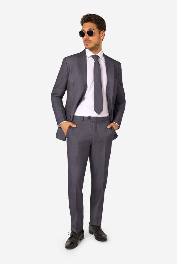 Een man draagt ​​een stijlvol antracietgrijs pak met een wit overhemd en ziet er zelfverzekerd uit. Perfecte formele outfit voor speciale gelegenheden, bruiloften en feesten. Hij kijkt zijwaarts. Volledig beeld en draagt zonnebril.