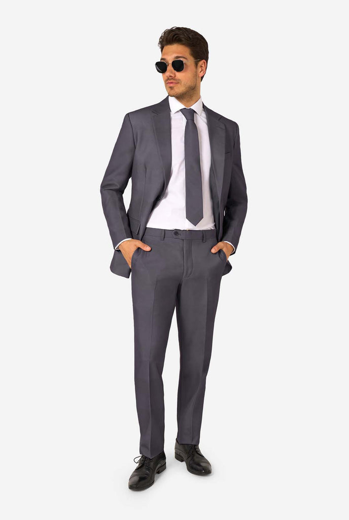 Een man draagt ​​een stijlvol antracietgrijs pak met een wit overhemd en ziet er zelfverzekerd uit. Perfecte formele outfit voor speciale gelegenheden, bruiloften en feesten. Hij kijkt zijwaarts. Volledig beeld en draagt zonnebril.