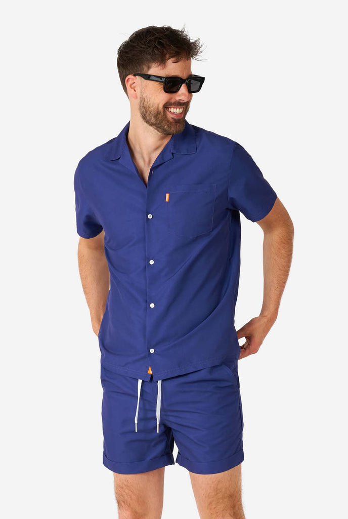 Man met blauwe zomerset bestaande uit shirt en korte broek en zonnebril.