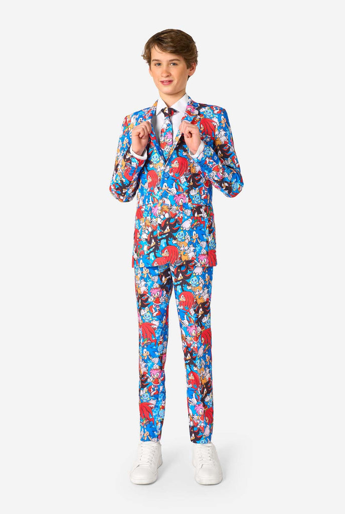 Tienerjongen draagt ​​een opvallend Sonic-tienerpak met kleurrijke prints van Sonic the Hedgehog-personages, waaronder Sonic, Knuckles, Tails en Amy, gecombineerd met een bijpassende stropdas en een wit overhemd. Volledig lichaam.