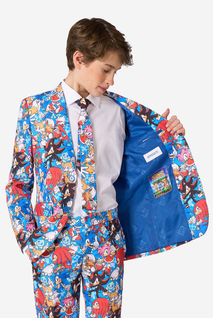 Tienerjongen draagt ​​een opvallend Sonic-tienerpak met kleurrijke prints van Sonic the Hedgehog-personages, waaronder Sonic, Knuckles, Tails en Amy, gecombineerd met een bijpassende stropdas en een wit overhemd. Houd een kant van het jasje open.