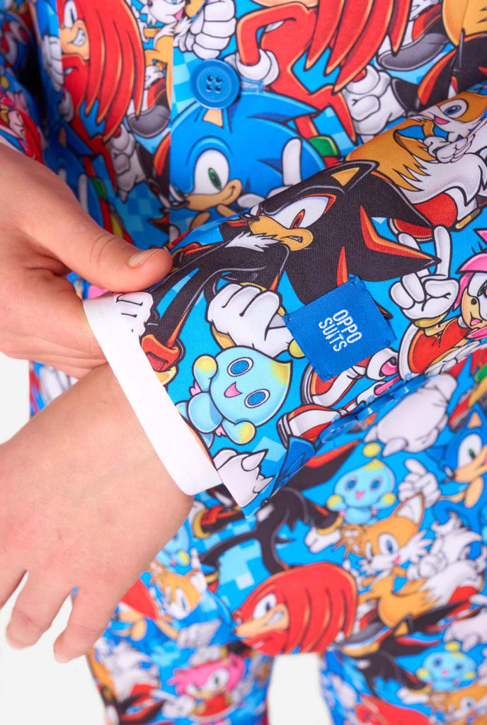 Tienerjongen draagt ​​een opvallend Sonic-tienerpak met kleurrijke prints van Sonic the Hedgehog-personages, waaronder Sonic, Knuckles, Tails en Amy, gecombineerd met een bijpassende stropdas en een wit overhemd. Close up van de mouwen.