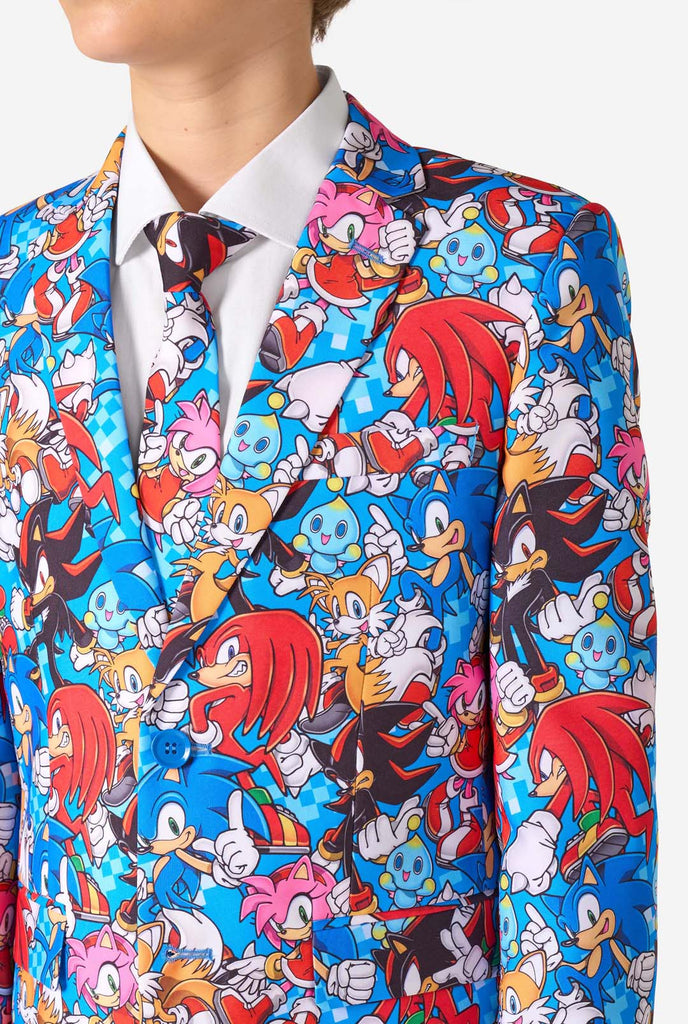 Tienerjongen draagt ​​een opvallend Sonic-tienerpak met kleurrijke prints van Sonic the Hedgehog-personages, waaronder Sonic, Knuckles, Tails en Amy, gecombineerd met een bijpassende stropdas en een wit overhemd. Close up van de borst.