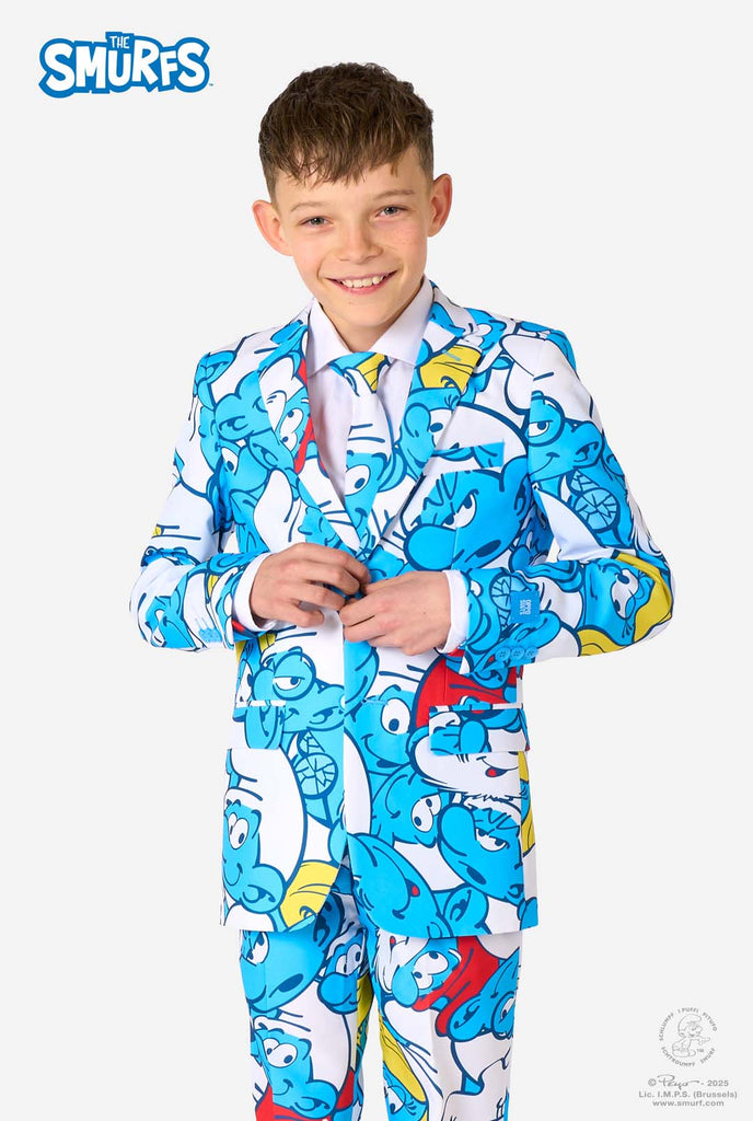 Een tienerjongen draagt ​​een opvallend Smurfenpak met kleurrijke cartoonfiguurprints in de kleuren blauw, rood, geel en wit, gecombineerd met een bijpassende stropdas en een wit overhemd, lachend tegen een effen achtergrond.