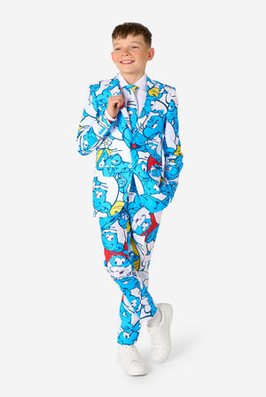 Een tienerjongen draagt ​​een opvallend Smurfenpak met kleurrijke cartoonfiguurprints in de kleuren blauw, rood, geel en wit, gecombineerd met een bijpassende stropdas en een wit overhemd, lachend tegen een effen achtergrond. Volledig lichaam.