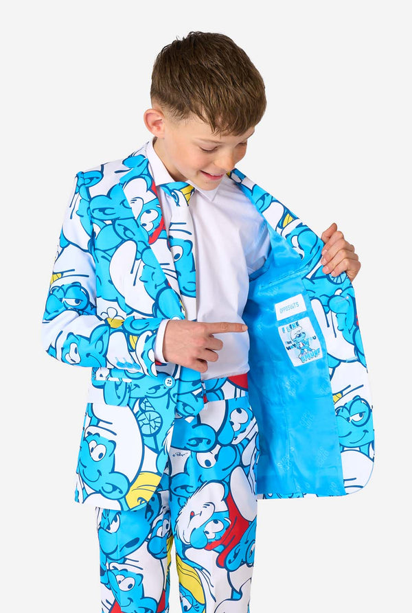 Een tienerjongen draagt ​​een opvallend Smurfenpak met kleurrijke cartoonfiguurprints in de kleuren blauw, rood, geel en wit, gecombineerd met een bijpassende stropdas en een wit overhemd, lachend tegen een effen achtergrond. Houd een kant van het jasje open.